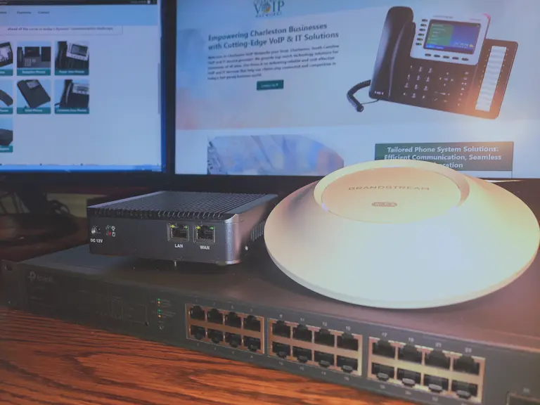Front view of a router, managed switch, and WiFi access point, with the Charleston VoIP Networks website displayed in the background.